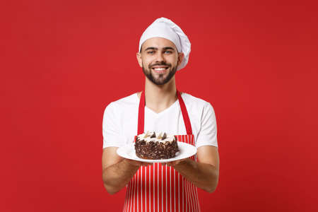 Smiling young bearded male chef cook or baker man in striped apron white t-shirt toque chefs hat posing isolated on red background. Cooking food concept. Mock up copy space. Holding plate with cake.の写真素材