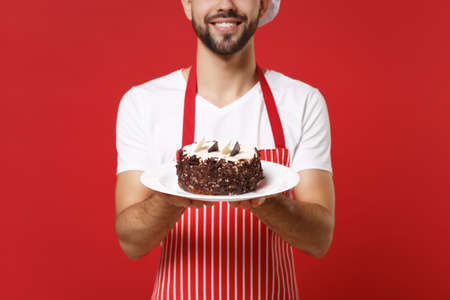 Cropped image of bearded male chef cook or baker man in striped apron white t-shirt posing isolated on red background, studio portrait. Cooking food concept. Mock up copy space. Hold plate with cake.の写真素材