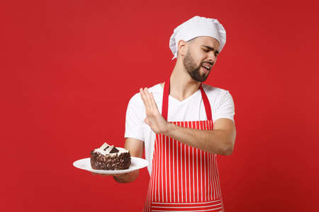 Bearded male chef cook or baker man in striped apron white t-shirt toque chefs hat posing isolated on red background. Cooking food concept. Mock up copy space. Showing stop gesture to plate with cake.の写真素材