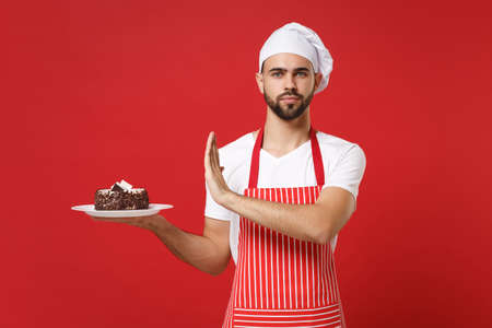 Bearded male chef cook or baker man in striped apron white t-shirt toque chefs hat posing isolated on red background. Cooking food concept. Mock up copy space. Showing stop gesture to plate with cake.の写真素材