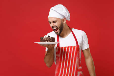 Crazy young bearded male chef cook or baker man in striped apron white t-shirt toque chefs hat posing isolated on red background. Cooking food concept. Mock up copy space. Holding plate with cake.の写真素材
