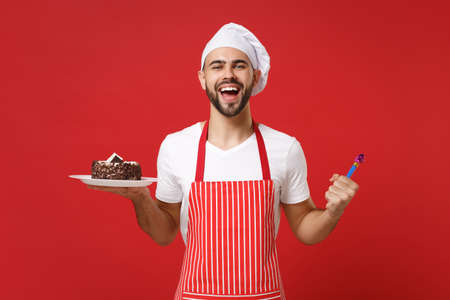 Cheerful male chef cook or baker man in striped apron white t-shirt toque chefs hat posing isolated on red background. Cooking food concept. Mock up copy space. Holding plate with cake, birthday pipe.の写真素材