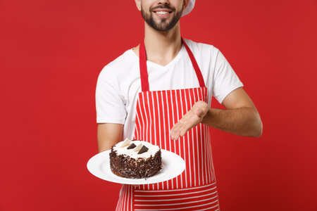 Cropped image of male chef cook or baker man in striped apron white t-shirt posing isolated on red background studio portrait. Cooking food concept. Mock up copy space. Point hand on plate with cake.の写真素材