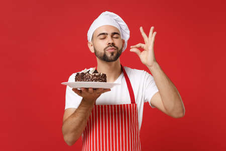 Bearded male chef cook or baker man in striped apron toque chefs hat posing isolated on red background. Cooking food concept. Mock up copy space. Hold plate with cake, making okay taste delight sign.の写真素材