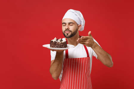 Male chef cook or baker man in striped apron toque chefs hat posing isolated on red background. Cooking food concept. Mock up copy space. Hold plate with cake, raised hand to face feeling food smell.の写真素材