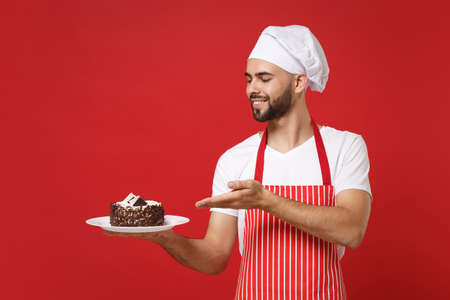Young bearded male chef cook or baker man in striped apron white t-shirt toque chefs hat posing isolated on red background. Cooking food concept. Mock up copy space. Pointing hand on plate with cake.の写真素材