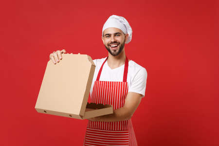 Funny young bearded male chef cook or baker man in striped apron toque chefs hat posing isolated on red background. Cooking food concept. Mock up copy space. Hold italian pizza in cardboard flatbox.の写真素材