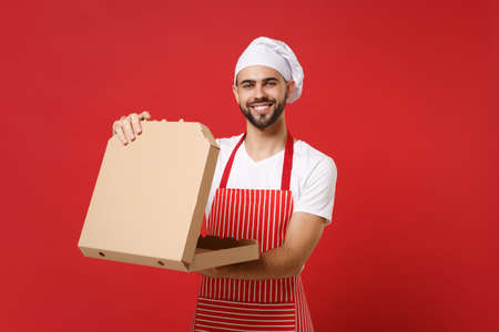 Smiling young bearded male chef cook or baker man in striped apron toque chefs hat posing isolated on red background. Cooking food concept. Mock up copy space. Hold italian pizza in cardboard flatbox.の写真素材