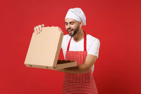 Curious young bearded male chef cook or baker man in striped apron toque chefs hat posing isolated on red background. Cooking food concept. Mock up copy space. Hold italian pizza in cardboard flatbox.の写真素材