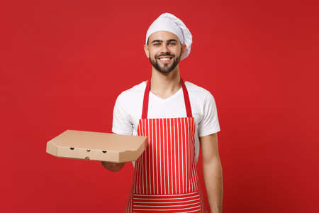 Smiling young bearded male chef cook or baker man in striped apron toque chefs hat posing isolated on red background. Cooking food concept. Mock up copy space. Hold italian pizza in cardboard flatbox.の写真素材