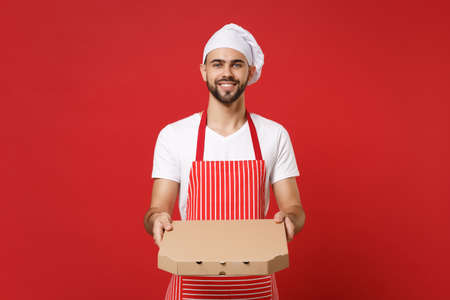 Smiling young bearded male chef cook or baker man in striped apron toque chefs hat posing isolated on red background. Cooking food concept. Mock up copy space. Hold italian pizza in cardboard flatbox.の写真素材