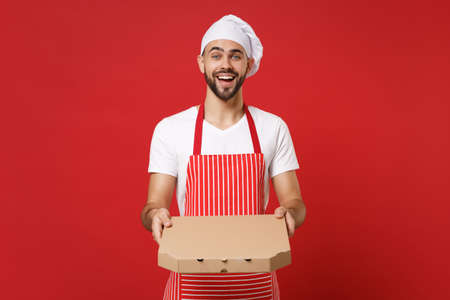 Joyful young bearded male chef cook or baker man in striped apron toque chefs hat posing isolated on red background. Cooking food concept. Mock up copy space. Hold italian pizza in cardboard flatbox.の写真素材