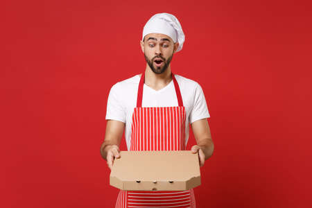 Shocked young bearded male chef cook or baker man in striped apron toque chefs hat posing isolated on red background. Cooking food concept. Mock up copy space. Hold italian pizza in cardboard flatbox.の写真素材