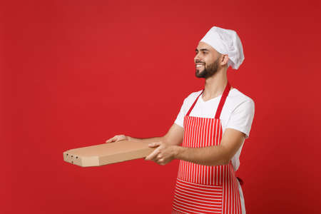 Smiling young bearded male chef cook or baker man in striped apron toque chefs hat posing isolated on red background. Cooking food concept. Mock up copy space. Hold italian pizza in cardboard flatbox.の写真素材