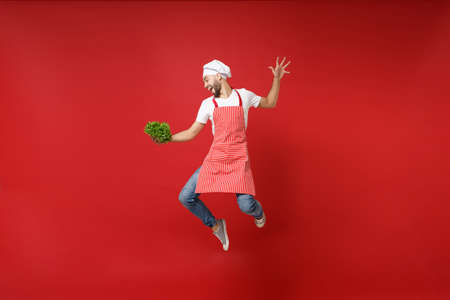 Funny young chef cook or baker man in striped apron toque chefs hat isolated on red background. Cooking food concept. Mock up copy space. Jumping, holding green fresh salad leaves, fooling around.の写真素材