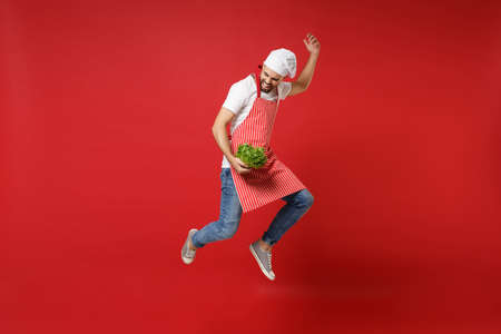 Funny young chef cook or baker man in striped apron toque chefs hat isolated on red background. Cooking food concept. Mock up copy space. Jumping, holding green fresh salad leaves, fooling around.の写真素材