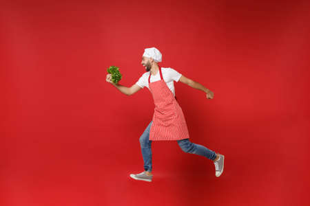 Crazy young chef cook or baker man in striped apron toque chefs hat isolated on red background. Cooking food concept. Mock up copy space. Jumping, holding green fresh salad leaves, fooling around.の写真素材