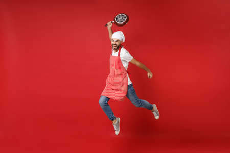 Crazy young bearded male chef cook or baker man in striped apron white t-shirt toque chefs hat posing isolated on red background. Cooking food concept. Mock up copy space. Jumping, holding frying pan.の写真素材