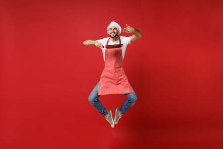 Smiling young bearded male chef cook or baker man in striped apron white t-shirt toque chefs hat posing isolated on red background. Cooking food concept. Mock up copy space. Jumping, hold frying pan.の写真素材