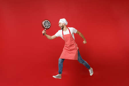 Crazy young bearded male chef cook or baker man in striped apron white t-shirt toque chefs hat posing isolated on red background. Cooking food concept. Mock up copy space. Jumping, holding frying pan.の写真素材