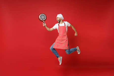 Funny young bearded male chef cook or baker man in striped apron white t-shirt toque chefs hat posing isolated on red background. Cooking food concept. Mock up copy space. Jumping, holding frying pan.の写真素材