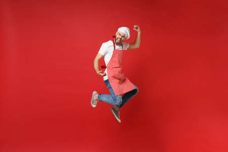 Screaming young male chef cook or baker man in striped apron white t-shirt toque chefs hat posing isolated on red background. Cooking food concept. Mock up copy space. Jump fooling around, having fun.の写真素材