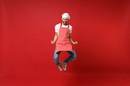 Happy joyful young male chef cook or baker man in striped apron white t-shirt toque chefs hat posing isolated on red background. Cooking food concept. Mock up copy space. Jumping doing winner gesture.の写真素材