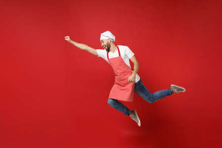 Crazy young male chef cook or baker man in striped apron white t-shirt toque chefs hat posing isolated on red background. Cooking food concept. Mock up copy space. Jumping, fooling around, having fun.の写真素材