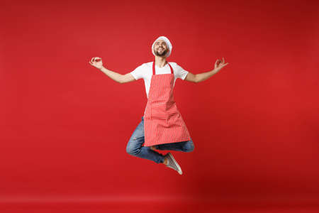 Smiling young male chef cook or baker man in striped apron toque chefs hat posing isolated on red background. Cooking food concept. Mock up copy space. Jumping, having fun, hold hands in yoga gesture.の写真素材
