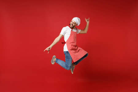 Crazy young male chef cook or baker man in striped apron toque chefs hat posing isolated on red background. Cooking food concept. Mock up copy space. Jumping, having fun, showing horns up gesture.の写真素材