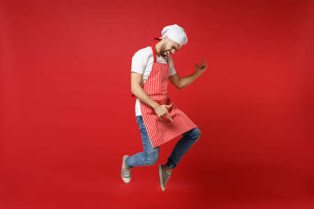 Crazy young male chef cook or baker man in striped apron white t-shirt toque chefs hat posing isolated on red background. Cooking food concept. Mock up copy space. Jumping, fooling around, having fun.の写真素材
