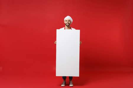 Smiling young chef cook or baker man in striped apron toque chefs hat isolated on red background. Cooking food concept. Mock up copy space. Holding white empty blank billboard place for text or image.の写真素材