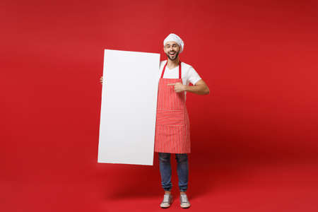 Excited chef cook or baker man in striped apron toque chefs hat isolated on red background. Cooking food concept. Mock up copy space. Pointing finger on white empty billboard place for text or image.の写真素材