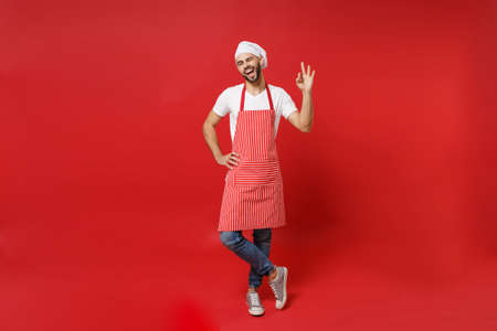 Funny young bearded male chef cook or baker man in striped apron white t-shirt toque chefs hat posing isolated on red background. Cooking food concept. Mock up copy space. Showing OK gesture blinking.の写真素材