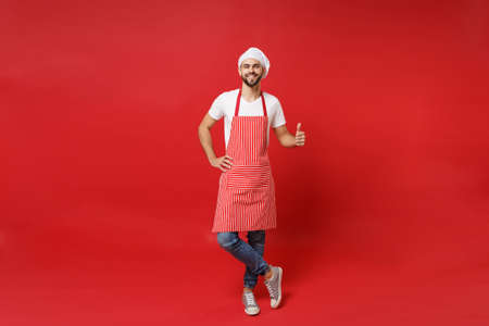 Smiling young bearded male chef cook or baker man in striped apron white t-shirt toque chefs hat posing isolated on red wall background. Cooking food concept. Mock up copy space. Showing thumb up.の写真素材