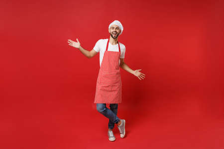 Cheerful young bearded male chef cook or baker man in striped apron white t-shirt toque chefs hat posing isolated on red wall background. Cooking food concept. Mock up copy space. Spreading hands.の写真素材