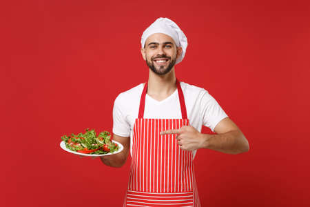 Smiling male chef cook or baker man in striped apron toque chefs hat posing isolated on red background. Cooking food concept. Mock up copy space. Pointing index finger on plate with vegetable salad.の写真素材