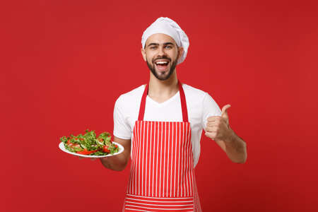 Joyful young male chef cook or baker man in striped apron toque chefs hat posing isolated on red background. Cooking food concept. Mock up copy space. Hold plate with vegetable salad showing thumb up.の写真素材