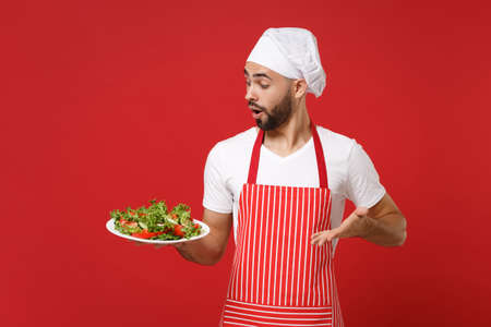 Shocked young bearded male chef cook or baker man in striped apron toque chefs hat posing isolated on red wall background. Cooking food concept. Mock up copy space. Hold plate with vegetable salad.の写真素材