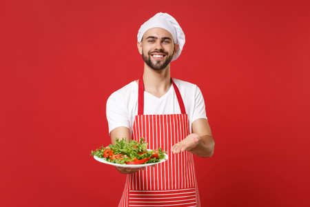 Smiling bearded male chef cook or baker man in striped apron toque chefs hat posing isolated on red background. Cooking food concept. Mock up copy space. Pointing hand on plate with vegetable salad.の写真素材