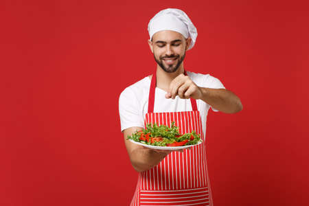Young bearded male chef cook or baker man in striped apron toque chefs hat posing isolated on red background. Cooking food concept. Mock up copy space. Holding plate, adds salt to vegetable salad.の写真素材