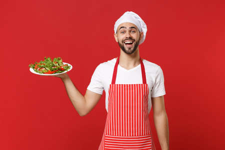Excited young bearded male chef cook or baker man in striped apron toque chefs hat posing isolated on red wall background. Cooking food concept. Mock up copy space. Holding plate with vegetable salad.の写真素材