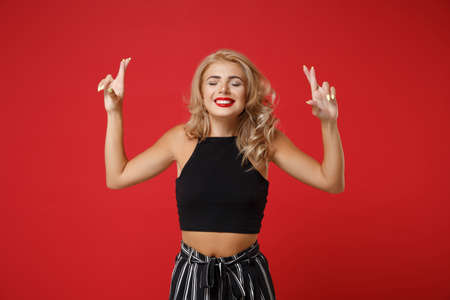 Young woman girl in black clothes posing isolated on red wall background. People lifestyle concept. Mock up copy space. Waiting for special moment, keeping fingers crossed, eyes closed, making wish.の写真素材