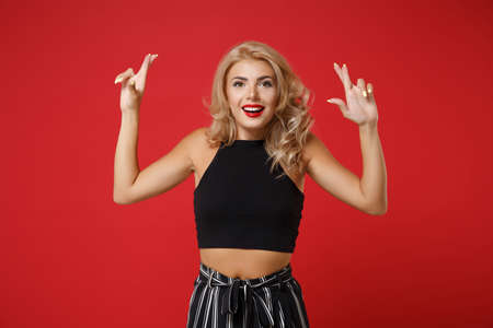 Excited young woman girl in black clothes posing isolated on bright red wall background. People lifestyle concept. Mock up copy space. Waiting for special moment, keeping fingers crossed, making wish.の写真素材