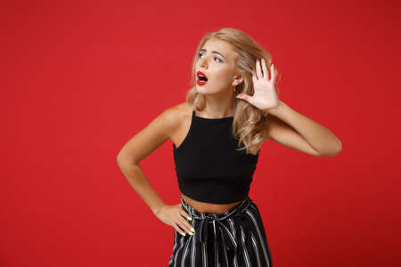 Curious young woman girl in black clothes posing isolated on red wall background, studio portrait. People sincere emotions lifestyle concept. Mock up copy space. Try to hear you with hand near ear.の写真素材