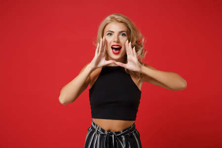Excited young woman girl in black clothes posing isolated on red wall background studio portrait. People sincere emotions lifestyle concept. Mock up copy space. Screaming with hand gesture near mouth.の写真素材