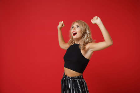 Excited young woman girl in black clothes posing isolated on red background studio portrait. People sincere emotions lifestyle concept. Mock up copy space. Looking aside, clenching fists like winner.の写真素材