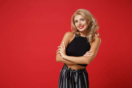 Smiling pretty young woman girl in black clothes posing isolated on bright red wall background studio portrait. People sincere emotions lifestyle concept. Mock up copy space. Holding hands crossed.の写真素材
