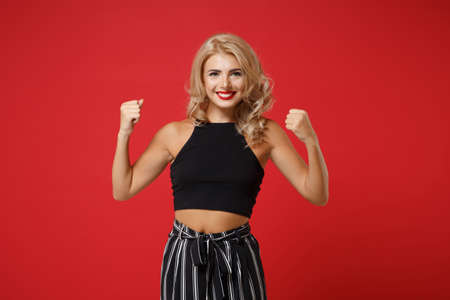 Cheerful young woman girl in black clothes posing isolated on bright red wall background studio portrait. People sincere emotions lifestyle concept. Mock up copy space. Clenching fists like winner.の写真素材