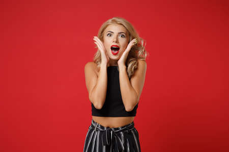 Shocked young woman girl in black clothes posing isolated on red background studio portrait. People sincere emotions lifestyle concept. Mock up copy space. Keeping mouth open, putting hands on cheeks.の写真素材